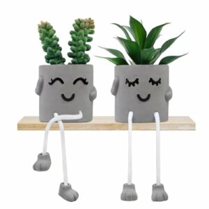 Dos macetas de cemento con caras sonrientes y patas colgantes, con plantas verdes artificiales.